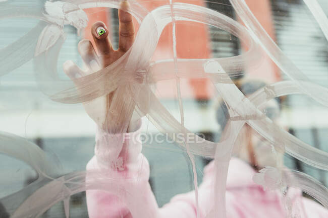 Durch das Glas eines bis zur Unkenntlichkeit ethnischen Typen in rosa Kapuzenpulli mit heller, kreativer Manikürmalerei auf transparentem Fenster mit Finger — Stockfoto
