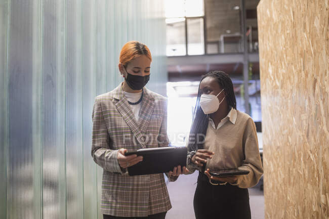 Багатоетнічні колеги-жінки з планшетами і в захисних масках, що стоять в коворкінгу і обговорюють проект — стокове фото