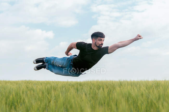 Hombre confiado en vaquero y camiseta extendiendo el puño hacia adelante mientras vuela por encima del suelo siendo superhéroe - foto de stock