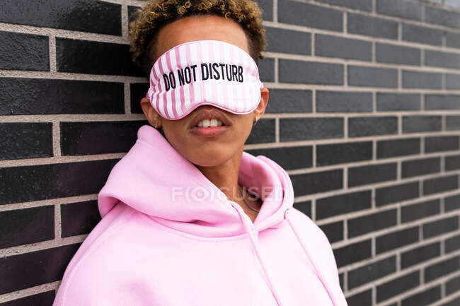 Непізнаваний молодий кучерявий етнічний хлопець в рожевому светрі і з зав'язаними очима з текстом Не турбуйтеся про цегляну стіну — стокове фото
