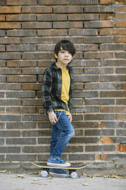 Vista lateral cuerpo completo de niño hispano étnico en ropa casual mirando a la cámara mientras está de pie en el monopatín en la pared de ladrillo en la calle - foto de stock