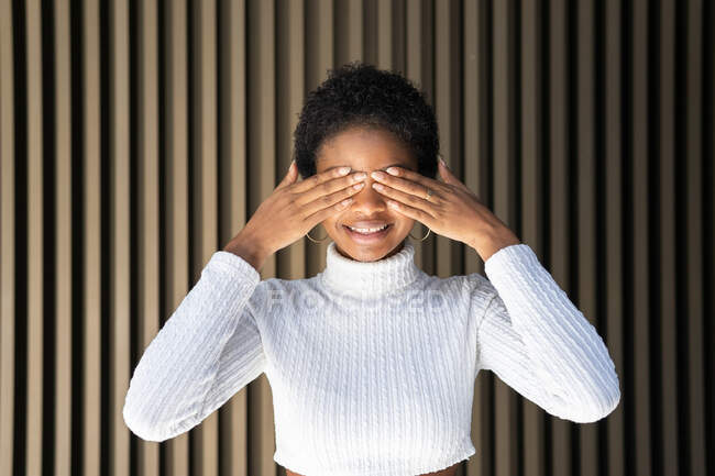 Mujer afroamericana feliz en suéter de moda cubriendo los ojos con sonrisa contra la pared rayada en la calle - foto de stock