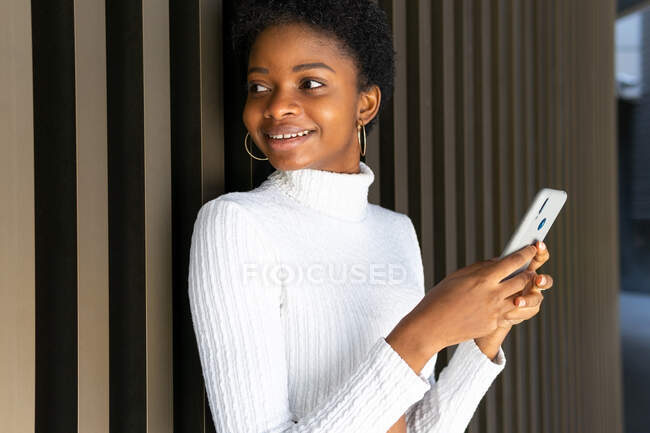 Feliz mujer joven y elegante negro en suéter de navegación en el teléfono inteligente, mientras que de pie cerca de la pared del edificio a rayas en la ciudad - foto de stock