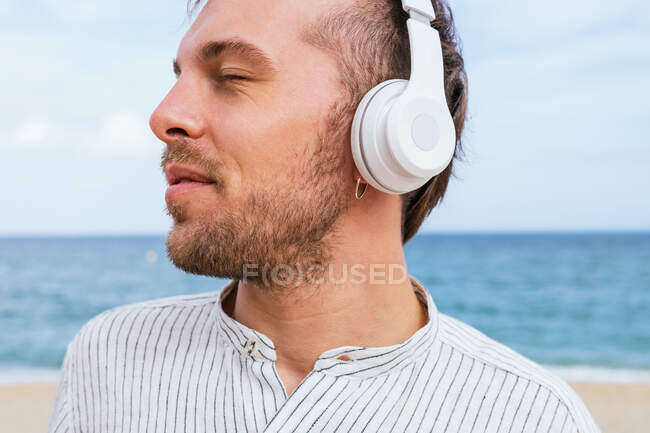 Giovane ragazzo barbuto spensierato in camicia casual elegante ascoltare musica attraverso cuffie wireless e godersi la brezza fresca mentre trascorre la giornata estiva sulla spiaggia di sabbia vicino al mare — Foto stock