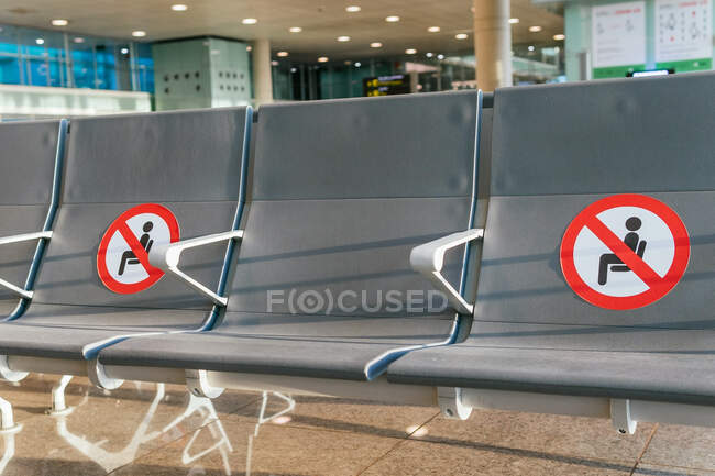 Fila de asientos vacíos con marcadores de restricción roja para el distanciamiento social en la sala de salidas del aeropuerto durante la epidemia de COVID - foto de stock