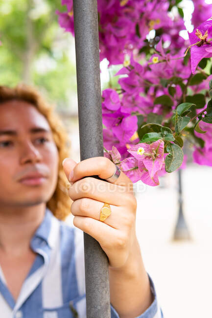 Модный мужчина с цветами в длинных волосах, шлифующий возле забора в парке в городе и смотрящий в сторону — стоковое фото