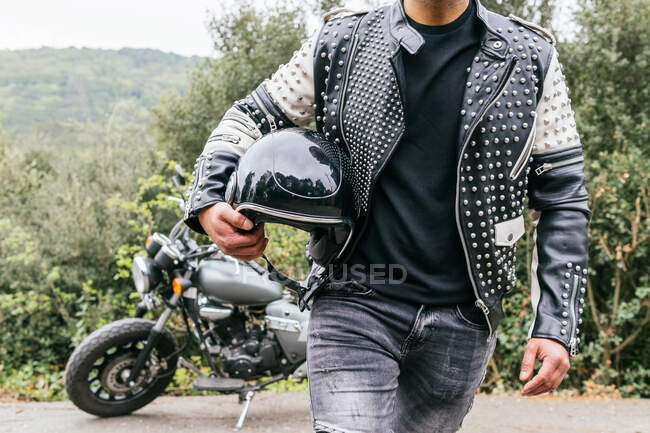 Анонимный мужчина-байкер в джинсах и кожаной куртке, держащий шлем в руке, стоя на асфальтовой дороге рядом с припаркованным современным мотоциклом — стоковое фото