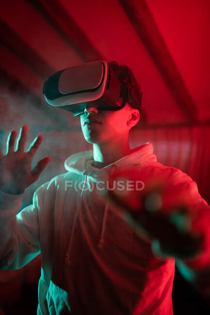 Man wearing hoodie while using VR goggles and standing in studio with red neon lights — Stock Photo