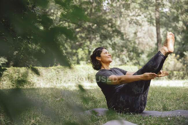Side view of content young ethnic female in sportswear performing Navasana pose and smiling while practicing yoga in green park — Stock Photo