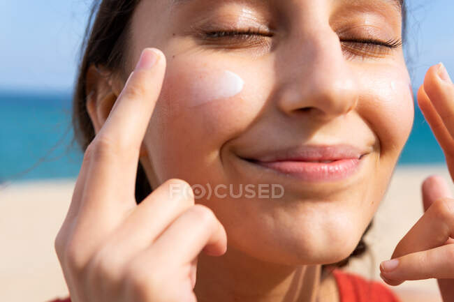 Улыбающаяся женщина с закрытыми глазами наносит лосьон для загара на лицо в солнечный день летом на пляже — стоковое фото