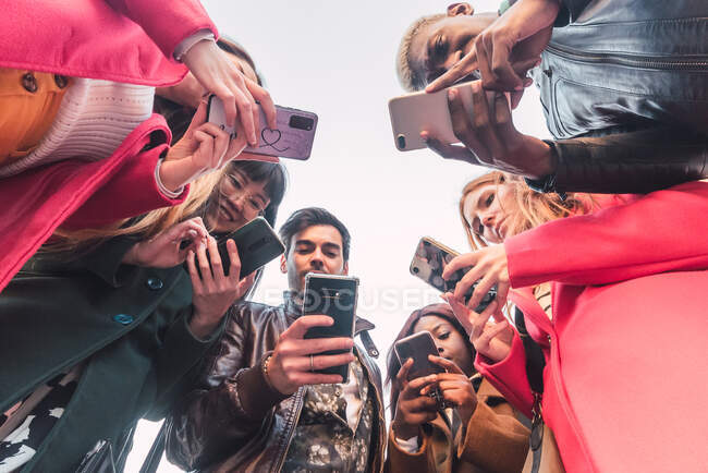 Знизу групи багаторасових друзів, що збираються в місті та переглядають мобільні телефони проти неба — стокове фото