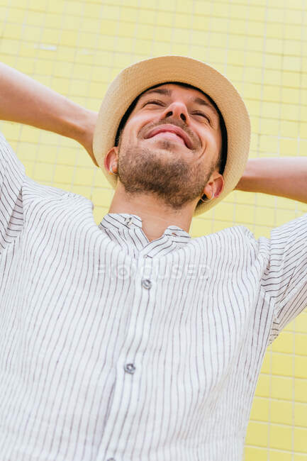 From below of happy young bearded hipster guy in trendy outfit and hat keeping hands behind head and smiling while chilling alone against yellow wall in summer day — Stock Photo