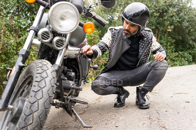 Full body of serious adult ethnic male biker in stylish outfit and helmet sitting on haunches and checking broken motorbike details on road near lush green trees in nature — Stock Photo