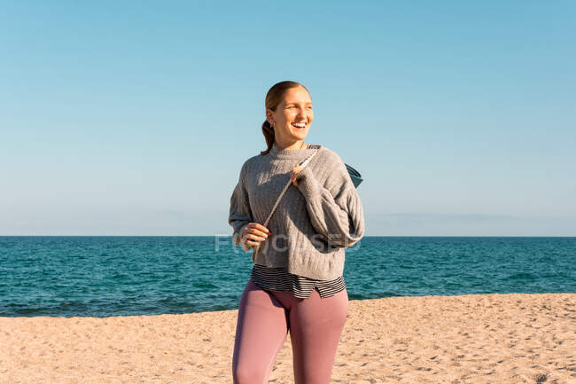 Joven hembra feliz en ropa deportiva con estera de yoga enrollada mirando a la distancia mientras está de pie en la playa de arena cerca del mar - foto de stock