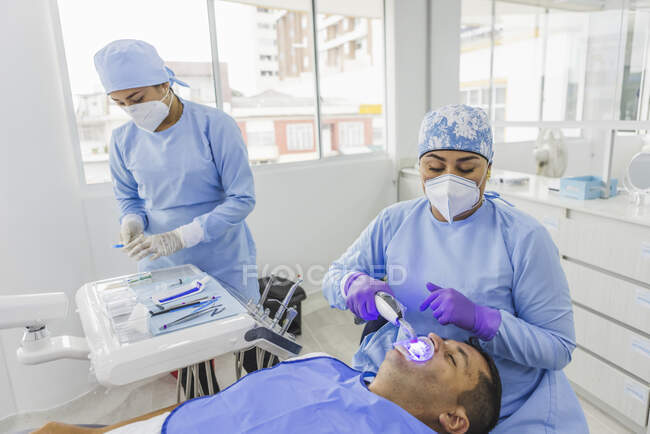 High angle of focused doctor wearing medical uniform treating client with dental tool with assistant preparing instruments in hospital — Stock Photo