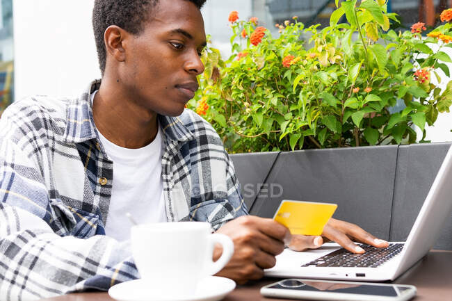 Hombre afroamericano enfocado que paga para la orden con tarjeta plástica mientras que usa el ordenador portátil durante compras en línea en la cafetería de la calle - foto de stock
