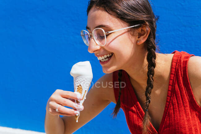Delighted female eating melting ice cream in waffle cone on sunny day in summer in street against blue wall — Stock Photo