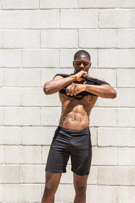 Confident African American male athlete with fit torso putting on tank top while standing near brick wall in city street on sunny day — Stock Photo