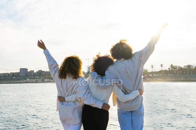 Back view of group of man and women with curly hair embracing each other with raising arms enjoying freedom while standing on seafront of city — Stock Photo