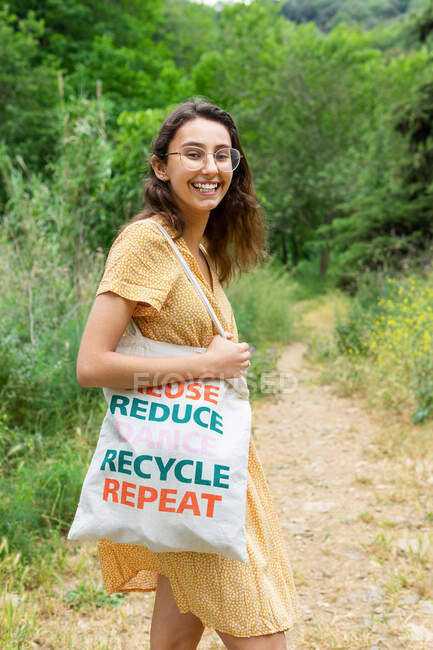 Seitenansicht der zufriedenen Frau, die mit umweltfreundlicher Stofftasche im Sommerpark steht und in die Kamera blickt — Stockfoto
