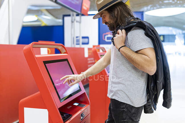 Junger Mann mit Bart und langen Haaren und Hut kauft Flugtickets am Automaten der Fluggesellschaft — Stockfoto