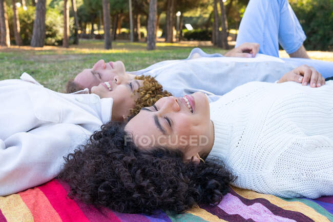 Diverse women and man with curly hair lying face to face on colorful plaid in park looking up — Stock Photo