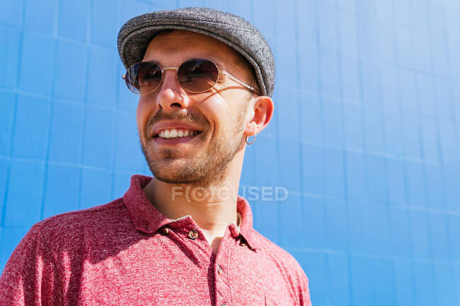 Молодий бородатий хлопець в повсякденній червоній сорочці поло і кепці дивиться на камеру над сонцезахисними окулярами і усміхнений дружній, стоячи на синій стіні на вулиці в літній день — стокове фото