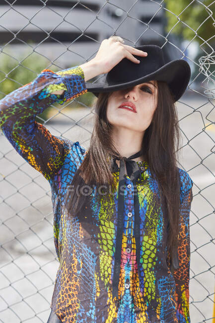 Mujer en ropa de moda de pie cerca de valla de malla en la ciudad en el día soleado y cubriendo los ojos con sombrero - foto de stock