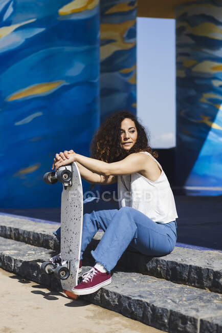 Полное тело вдумчивой женщины в повседневной одежде, сидящей на лестнице возле скейтборда на солнечной улице во время тренировки — стоковое фото
