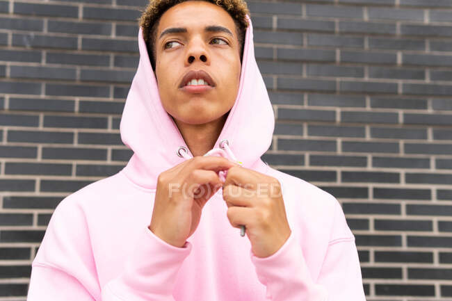 Fiducioso serio giovane ragazzo hipster dai capelli ricci in felpa rosa con cappuccio guardando lontano contro il muro di mattoni — Foto stock