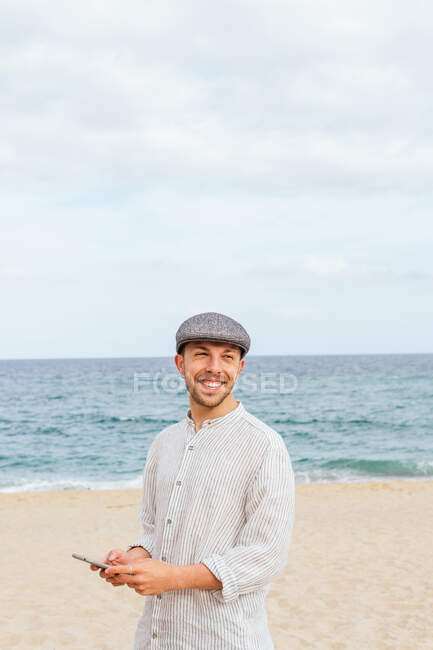 Молодой бородатый парень в стильной рубашке и кепке улыбается и смотрит в сторону, пока просматривает мобильный телефон на песчаном пляже у моря — стоковое фото