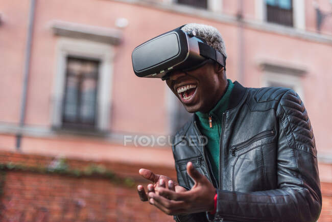 Excited African American male in VR goggles experiencing virtual reality and laughing in city street — Stock Photo
