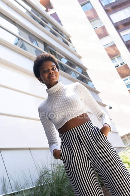 From below stylish self assured African American woman with hands on the pocket looking at camera while standing near modern apartment buildings on sunny day in city — Stock Photo