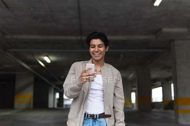 Giovane uomo latino sorridente in abiti casual utilizzando il telefono mentre in piedi nel parcheggio — Foto stock