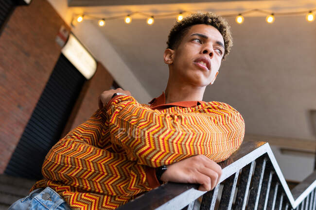 Von unten ein junger ethnischer Lockenkopf in stylischem, bunt gestreiftem Hemd, der sich an Geländer lehnt und nachdenklich wegschaut — Stockfoto