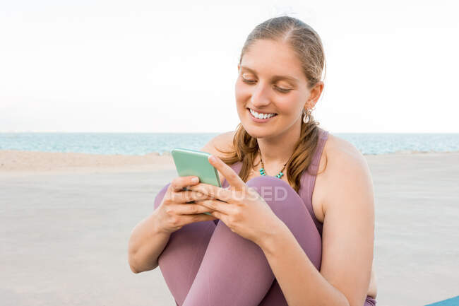Delighted female in activewear sitting on yoga mat browsing on mobile phone on seashore — Stock Photo