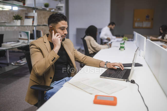 Empreendedor masculino étnico focado falando no telefone celular e trabalhando no local de trabalho enquanto se senta à mesa com laptop — Fotografia de Stock