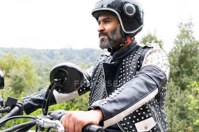 Biker di etnia maschile barbuto in giacca di pelle nera e casco in sella alla moto moderna su strada asfaltata tra rigogliosi alberi verdi che crescono nella valle montuosa — Foto stock