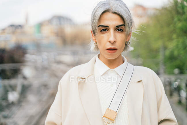 Самоуверенная трансгендерка в стильном пальто, стоящая на мосту и смотрящая в камеру — стоковое фото
