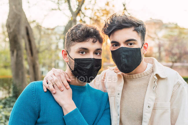 Люблячі ЛГБТ пари чоловіків носять захисні маски, обіймаючись в парку під час епідемії коронавірусу і дивлячись на камеру — стокове фото