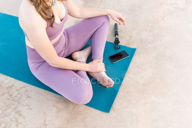 Сверху урожая анонимная женщина сидит в позе лотоса и практикует йогу на коврике с мобильного телефона — стоковое фото