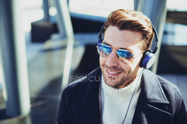 Maschio barbuto sorridente in occhiali da sole moderni ascoltare la canzone da cuffie in città in retroilluminazione — Foto stock
