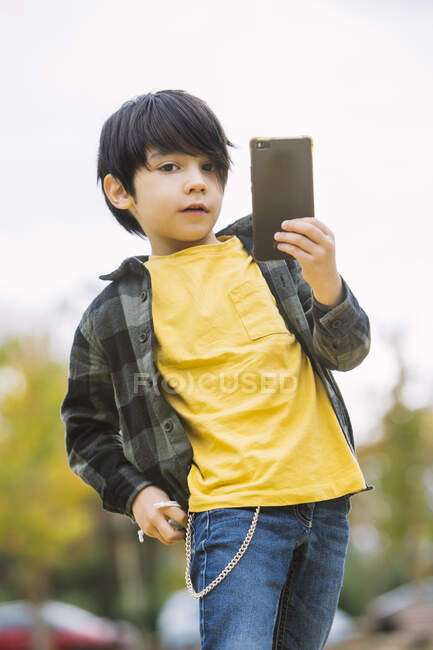 Очаровательный мальчик с черными волосами в повседневной одежде просматривает мобильный телефон и смотрит в камеру, стоя на городской улице на размытом фоне — стоковое фото