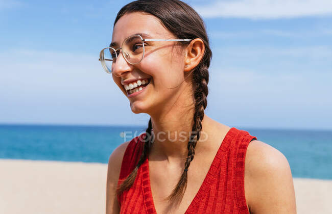 Mulher alegre em roupas de verão com tranças em pé na costa arenosa com mar azul calmo no dia ensolarado — Fotografia de Stock