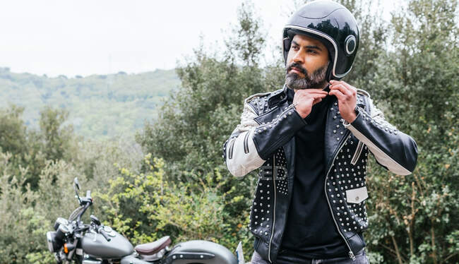Самоуверенный взрослый бородатый велосипедист в стильной кожаной куртке, регулирующий защитный шлем и смотрящий в сторону в природе — стоковое фото