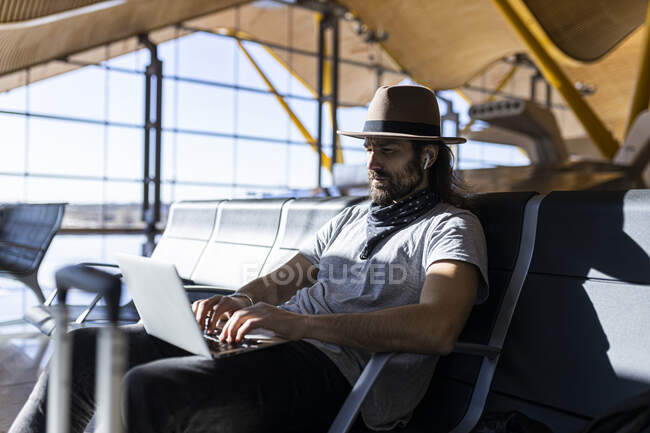 El tipo en el sombrero en el aeropuerto en la sala de espera sentado esperando su vuelo, con auriculares inalámbricos para escuchar música mientras trabaja con su computadora portátil - foto de stock