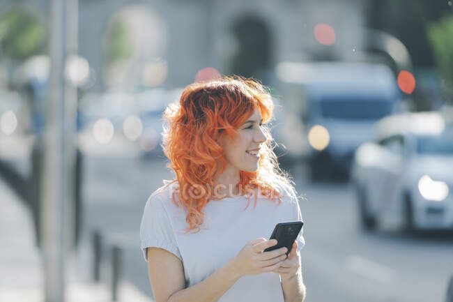 Cheerful redhead female on street and messaging on social media on mobile phone — Stock Photo