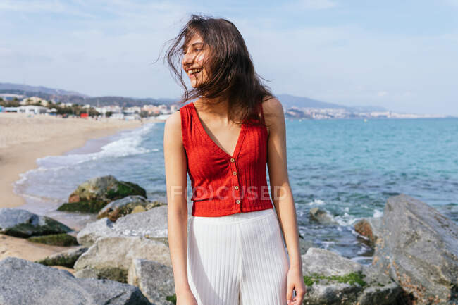 Mujer positiva en traje elegante de pie en piedra en la orilla del mar y disfrutar de un día soleado durante las vacaciones de verano - foto de stock