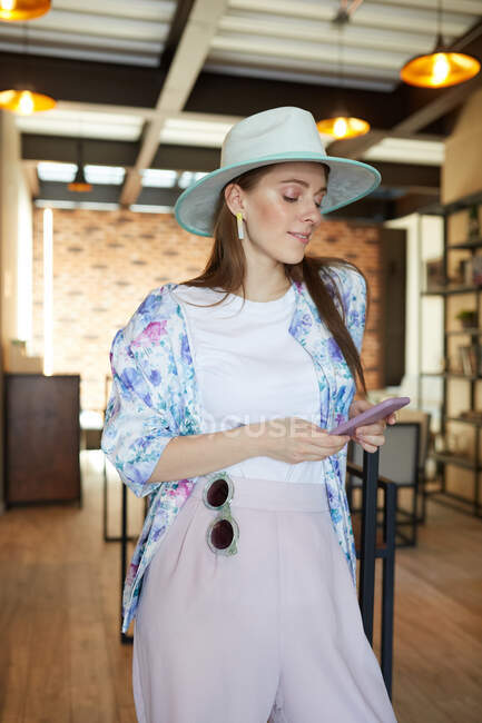 Усміхнена молода жінка в стильному одязі з мобільним телефоном в кафетерії з лампами — стокове фото