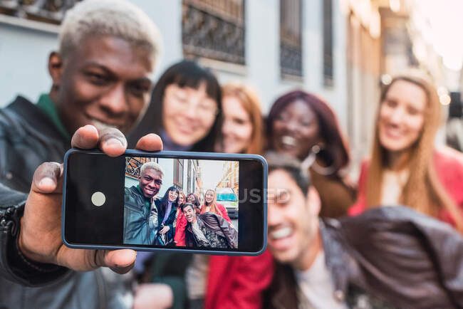 Афроамериканец делает селфи со смартфоном с компанией многорасовых друзей, стоящих на улице вместе — стоковое фото